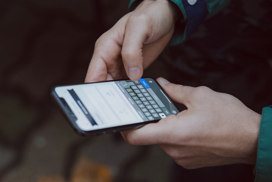 Close-up of a person holding a smartphone with both hands, using the touchscreen to input information or request a quote for house removal services. The person is wearing a dark green jacket with a hi