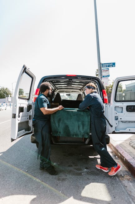 A person wearing a dark green shirt and dark trousers is seen during a home relocation process, holding a cardboard box with both hands. The box is sealed with brown packing tape, and a white and red 