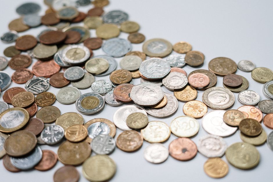 Close-up image showing a black calculator with blue and white buttons partially visible in the top left corner, placed next to three stacks of UK coins (pennies, two-pound, and one-pound coins) on a w