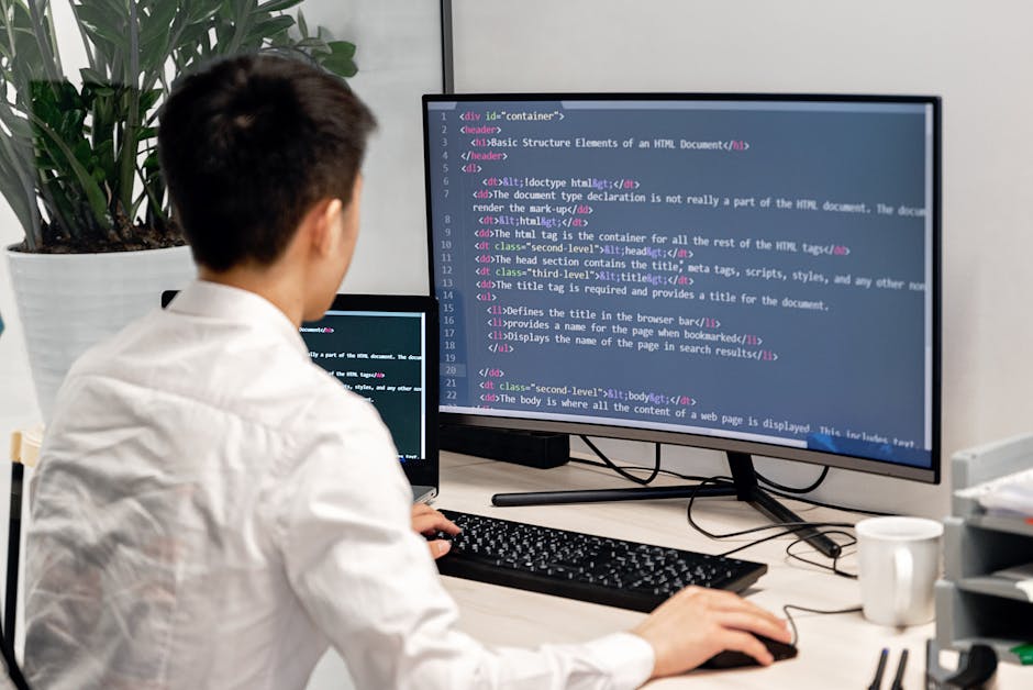 A person with short brown hair, dressed in a white shirt, working at a desk with multiple monitors. The main monitor displays a web development code editor with HTML code visible. An additional smalle